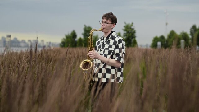 Caucasian teenage musician playing saxophone outdoors in urban park among tall grass in slow motion. Clip perfect for music education campaigns, arts festivals and youth talent promotions