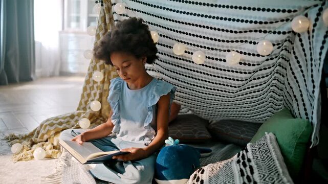 Preteen girl reading book cozy blanket fort. Fairy lights kids play tent home.