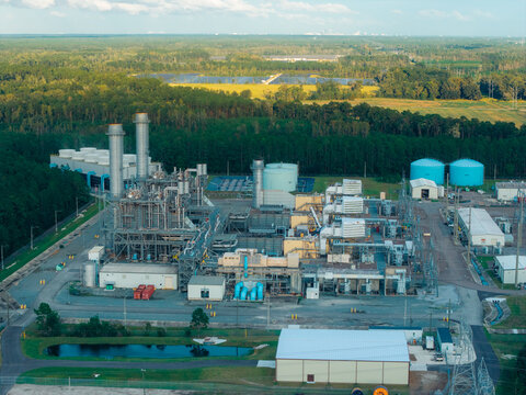 Aerial view of an industrial power plant facility featuring large cooling towers, storage tanks, and complex machinery surrounded by dense forest in Jacksonville, Florida, United States.