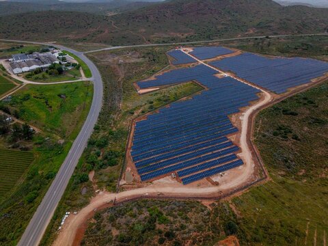 Aerial view of a large solar farm with rows of photovoltaic panels situated in a rural landscape near a road and buildings in Bonnievale, Western Cape, South Africa.