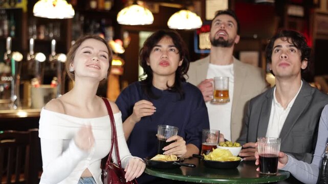 Group of emotional football fans watching match of favorite team at table in big sports bar, and screaming chants together 