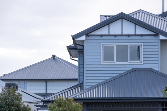 Roofline and upper floor of modern two-storey suburban houses in Australia, with horizontal weatherboard cladding, a gable with panels and trim, a window and corrugated metal hip roofs. 