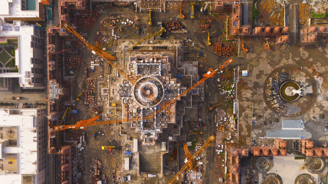 Aerial view of a large construction site featuring a central temple structure with multiple yellow cranes, scaffolding, and circular water features in Surat, Gujarat, India.