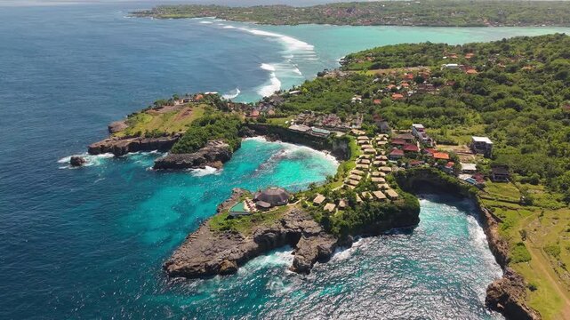 Waves crashing into a rocky cove surrounded by island vegetation and small resort huts along the coastline under bright sunlight