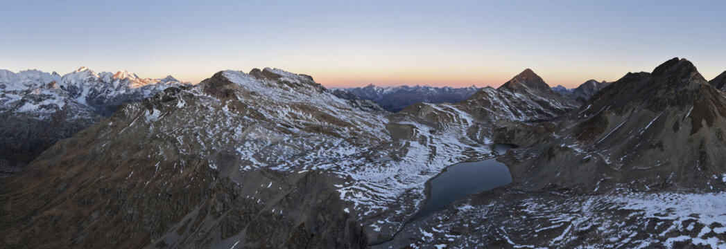 Aerial view of Engadin valley featuring snow-capped mountain peaks, a serene alpine lake, and rocky slopes illuminated by the soft light of golden hour, Grisons, Switzerland.