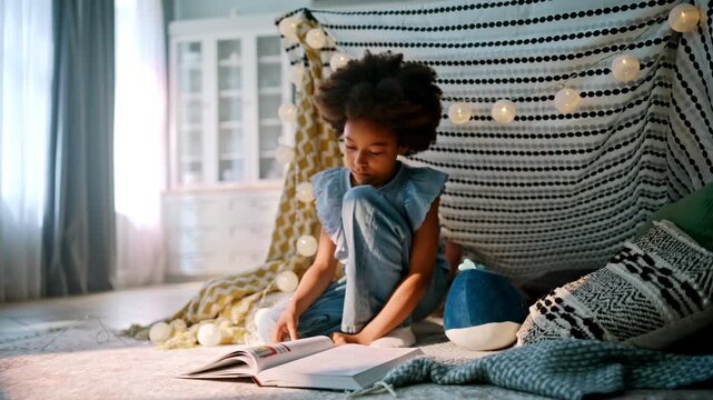 Schoolgirl reading book blanket fort. African american kid turning pages home.