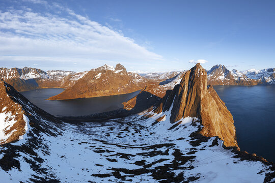 Aerial view of the dramatic Segla mountain peak rising above the deep blue fjord, surrounded by snow-covered slopes and rugged terrain under a clear sky in Senja, Troms, Norway.