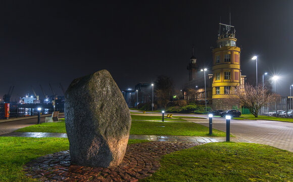 View of the Gdansk Harbour Master's Office illuminated at night with a large stone monument in the foreground, set in a park area in Gdansk, Pomeranian Voivodeship, Poland.