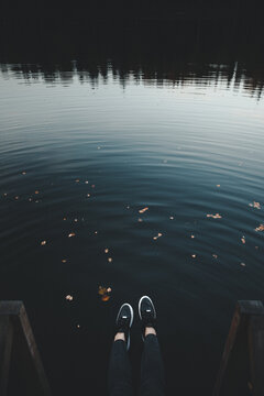 View of a person's legs and feet wearing black sneakers dangling over the calm, dark water of a lake with scattered autumn leaves in Neeruti, Laane-Viru County, Estonia.