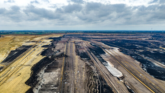 Aerial view of Garzweiler surface mine showing open-pit coal mining operations with industrial machinery and layered earth textures under a cloudy sky in Juchen, North Rhine-Westphalia, Germany.