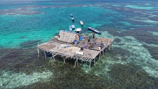 Aerial view of a traditional wooden stilt house platform surrounded by turquoise waters and coral reefs, with fishermen on the deck and boats nearby in Wakatobi, Southeast Sulawesi, Indonesia.