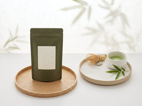 Matcha Green Tea Pouch Packaging with Blank Label on Wooden Tray with Tea Cup and Bamboo Leaves on White Table. Concept of Product Mockup, Japanese Tea, Organic Wellness, Branding, and Minimal Zen Sty