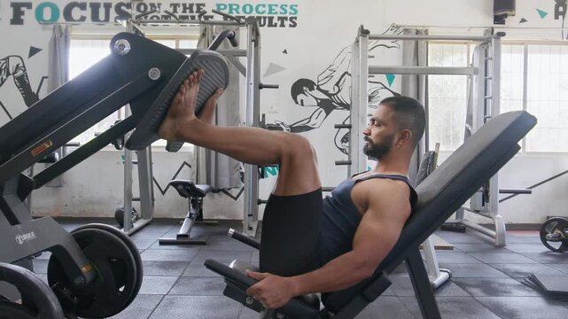 A young Indian Asian fit, muscular, confident male or man performs a leg press exercise during a resistance weight training workout in a modern gym. Fitness, health, bodybuilding, wellness concept.