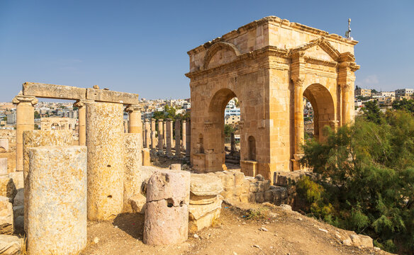 View of the ancient Arch of Hadrian surrounded by weathered stone columns and ruins under a clear blue sky in Jerash, Jerash Governorate, Jordan.