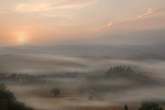 View of rolling hills covered in morning mist at sunrise with cypress trees and farmhouses in Tuscany, Italy.