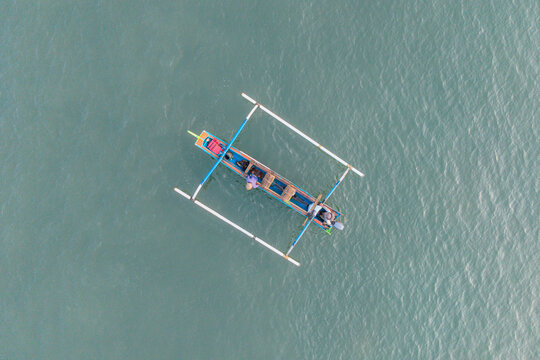 Top down aerial drone view of traditional blue and orange Indonesian jukung outrigger fishing boat floating on calm greenish ocean water with two fishermen on board