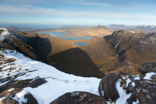 View of the rugged mountain landscape of Ben Mor Coigach with snow-covered rocky foreground, deep valleys, and distant lochs in Ullapool, Scotland, United Kingdom.