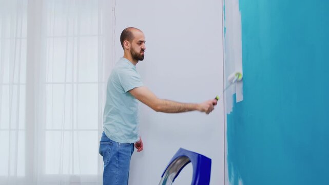 Man rolls turquoise paint across the middle of an apartment wall beside a step ladder and protected floor, combining interior painting, color change and hands-on DIY home renovation work.