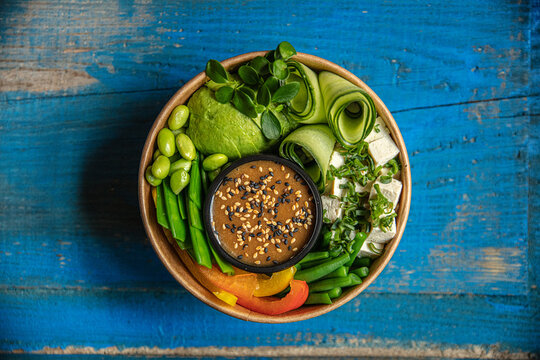 View of a healthy poke bowl with avocado, tofu, edamame, cucumber ribbons, and sesame dressing in a paper container on a blue wooden surface, Latvia.