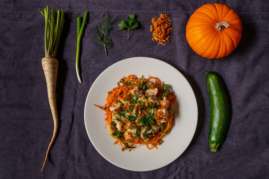 View of vegetable noodles with chicken, herbs, and fried onions, surrounded by raw ingredients including parsnip, pumpkin, and zucchini on a dark fabric background in Latvia.