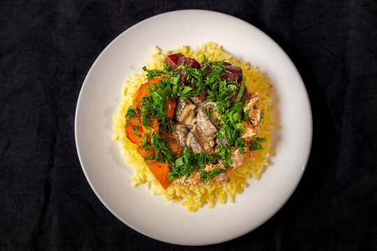 View of a plate of yellow rice topped with roasted carrots, mushrooms, salmon, and fresh green herbs, Latvia.