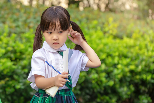Diligent Asian girl student sitting on a bench with her backpack, writing and learning outdoors.