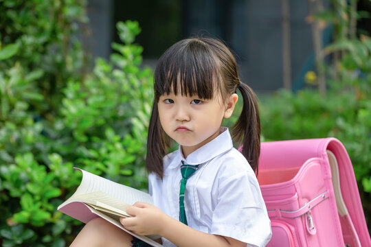 Diligent Asian girl student sitting on a bench with her backpack, writing and learning outdoors.