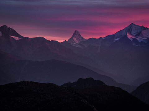 Aerial view of the iconic Matterhorn peak illuminated by the vibrant purple and pink hues of a sunset sky over the rugged alpine landscape of the Swiss Alps, Valais, Switzerland.