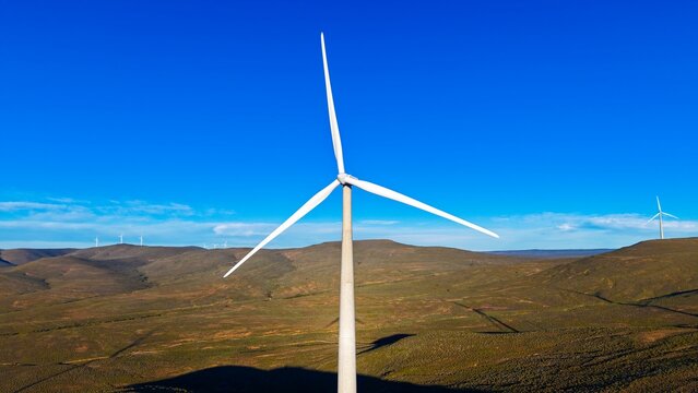 Aerial view of Roggeveld Wind Farm featuring a large wind turbine in the foreground, surrounded by rolling hills and arid landscape in Matjiesfontein, Western Cape, South Africa.