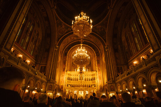 View of the ornate interior of the Timisoara Orthodox Metropolitan Cathedral with glowing chandeliers and religious iconography above a congregation in Timisoara, Timis, Romania.