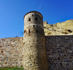 A low-angle view showcases a medieval stone watchtower and fortified defensive wall built against sandstone cliffs at the David Gareja monastery complex under a clear blue sky in southeastern Georgia © Gregory