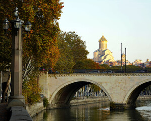 An ornate classical street lamp frames an arched stone bridge crossing the Kura (Mtkvari) River, with the Holy Trinity Cathedral of Tbilisi (Sameba) rising above the city skyline in golden late-aftern © Gregory