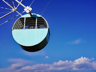 A vintage cylindrical turquoise Ferris wheel gondola hangs suspended against a vibrant blue sky dotted with soft white clouds in Chiatura, Georgia © Gregory
