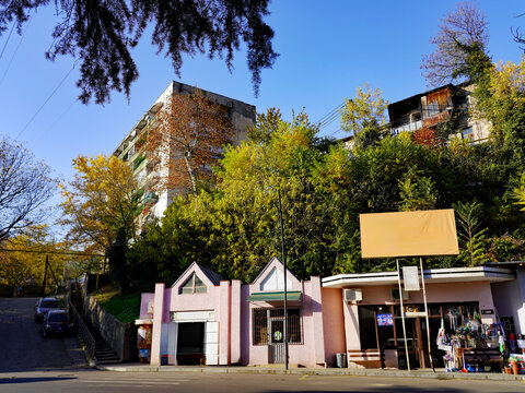 Pastel-pink street-level storefronts with a blank billboard stand beneath lush autumn trees and Soviet-era residential apartment blocks along an uphill road in Tbilisi, Georgia