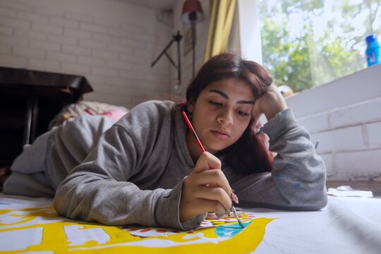 Young female artist lying on floor painting on large canvas in bright studio
