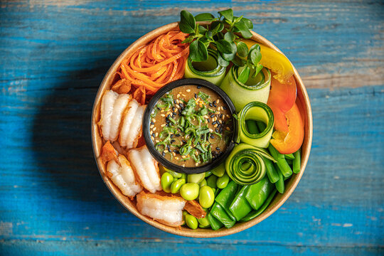 View of a fresh poke bowl with shrimp, edamame, cucumber rolls, shredded carrots, and bell peppers with a central sesame sauce in a paper bowl on a rustic blue wooden background in Latvia.