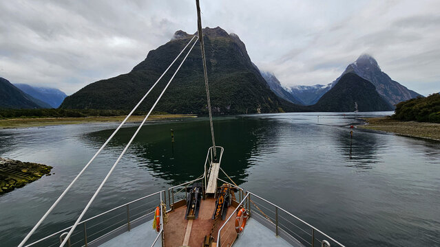 View of the bow of a boat sailing through the calm waters of Milford Sound Piopiotahi surrounded by towering mountains under a cloudy sky in Te Anau, Southland Region, New Zealand.