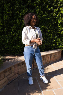 African woman standing in courtyard by bench and hedge laughing wearing shirt, jeans