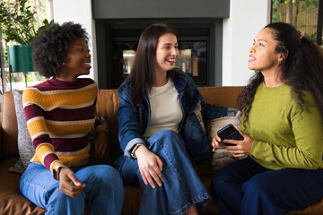 Fototapeta premium Diverse female friends chatting on leather couch by fireplace in living room holding smartphone