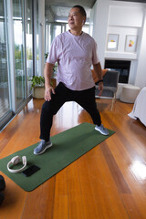Fototapeta premium Senior Asian man stretching on green mat at home wearing pink tee black pants headphones smartwatch