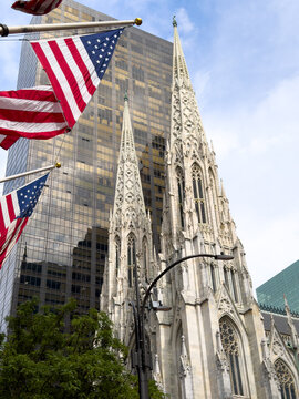 A Gothic cathedral stands beside towers on Fifth Avenue.. Red and white flag stripes cross the foreground above carved stone and pointed windows.