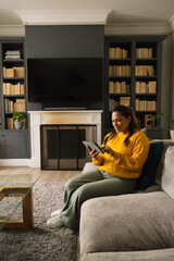 Fototapeta premium African American female sitting on sectional at home in mustard sweater using tablet, copy space