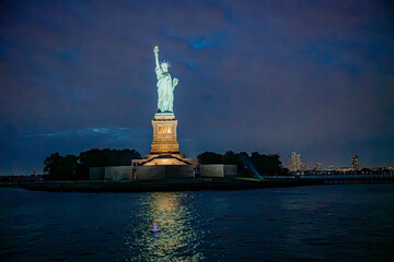 The Statue of Liberty rises above the island after dark.. The green figure and gold pedestal sit above rippling water with trees and distant lights to the right. © white78