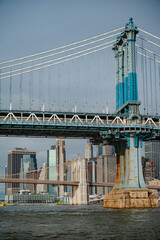 Fototapeta premium A Manhattan Bridge tower frames the skyline above the river.. Steel cables cross a vertical view above a brown stone pier with the Brooklyn Bridge in the distance.