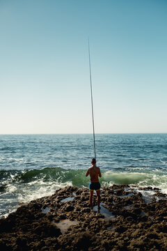 View of a man fishing with a long rod from the rocky shore of Praia da Bicas in Sines, Setubal, Portugal.