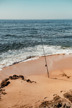 View of a fishing rod stuck in the sand on the shore of Praia da Bicas with gentle ocean waves rolling onto the beach in Sines, Setubal, Portugal.