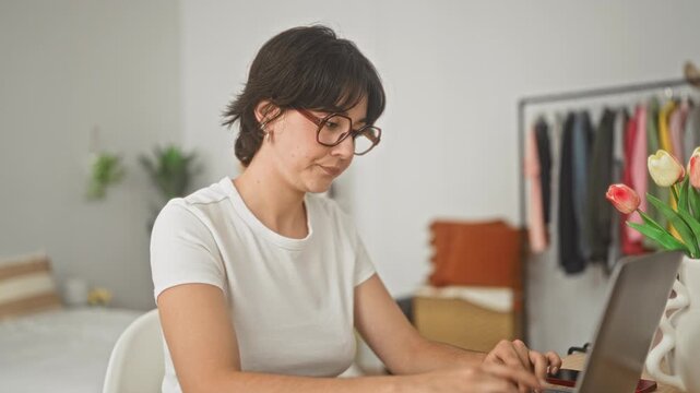 Woman adjusts glasses and types on laptop with hands near face at a studio desk beside a clothing rack and tulips; tiredness.