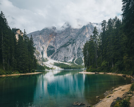 View of Pragser Wildsee with its turquoise water reflecting the towering, cloud-capped Dolomites mountains and surrounding evergreen pine forest in Prags, Trentino-Alto Adige, Italy.