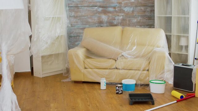 Sofa and nearby furniture are protected with plastic while paint buckets and tools are arranged for a living room renovation and interior makeover.
