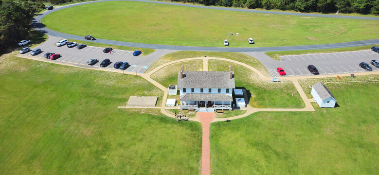 View of the historic Bodie Island Lighthouse Keeper's Quarters surrounded by green lawns, paved parking areas with cars, and walkways in Nags Head, North Carolina, United States.